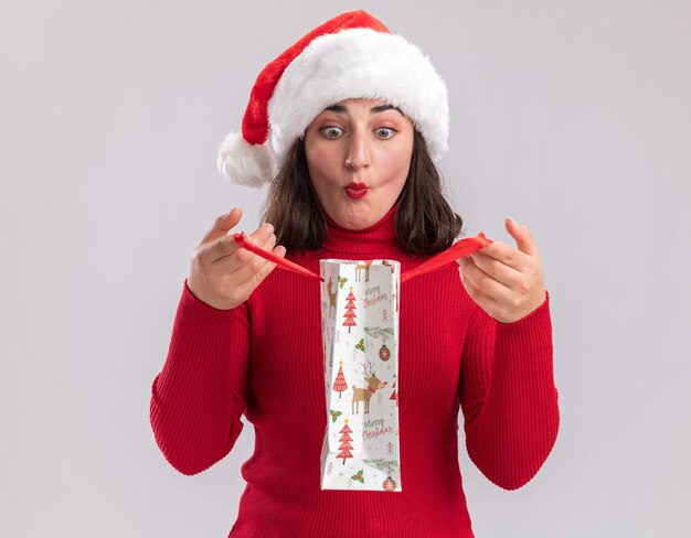 Photo gratuite jeune fille en pull rouge et santa hat holding sac en papier coloré avec des cadeaux de noël sac d'ouverture à l'intérieur surpris debout sur fond blanc