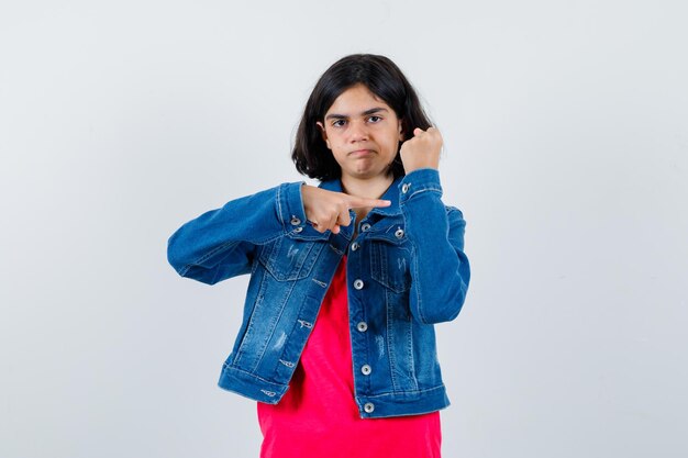 Jeune fille qui s'étend du doigt vers le bras en t-shirt rouge et veste en jean et a l'air sérieux. vue de face.