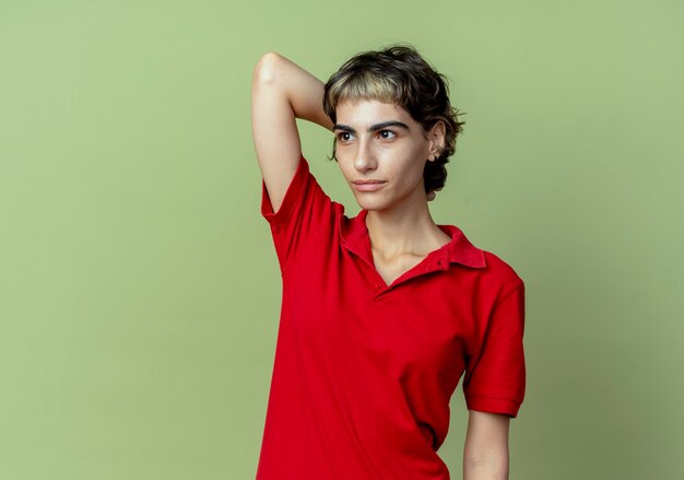 Jeune fille de race blanche avec coupe de cheveux de lutin à côté de mettre la main derrière la tête isolée sur fond vert olive avec espace de copie