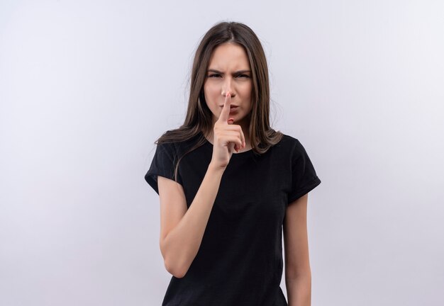 Jeune fille de race blanche portant un t-shirt noir montrant le geste de silence sur un mur blanc isolé