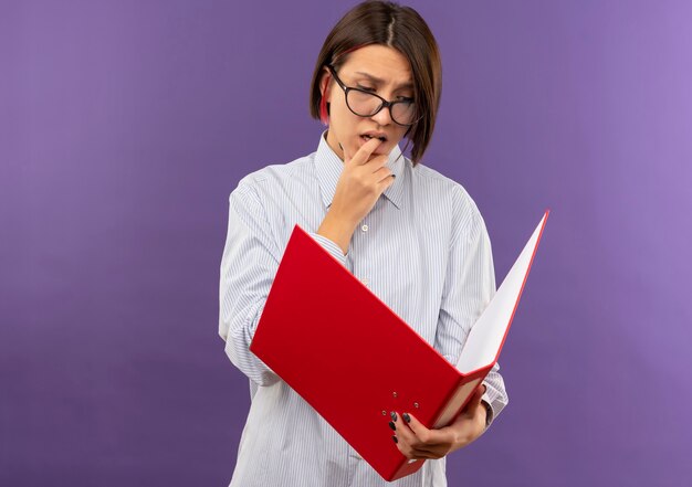 Jeune fille réfléchie de centre d'appels portant des lunettes tenant et regardant le dossier et se mordant le doigt isolé sur le mur violet