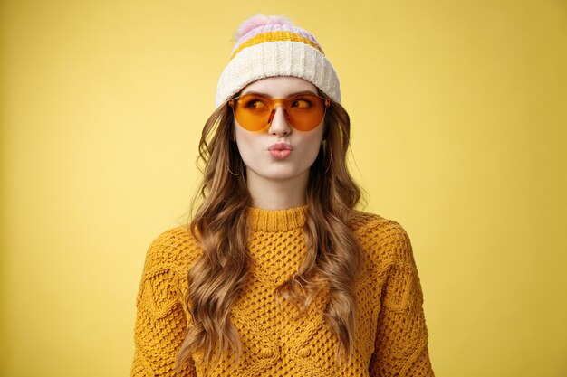 Jeune fille réfléchie glamour élégante faire des lèvres pliantes desicion pensant idiot en choisissant le produit détourner le regard vers la gauche portant des lunettes de soleil chapeau pull chaud profitant de la station de ski de vacances, fond jaune