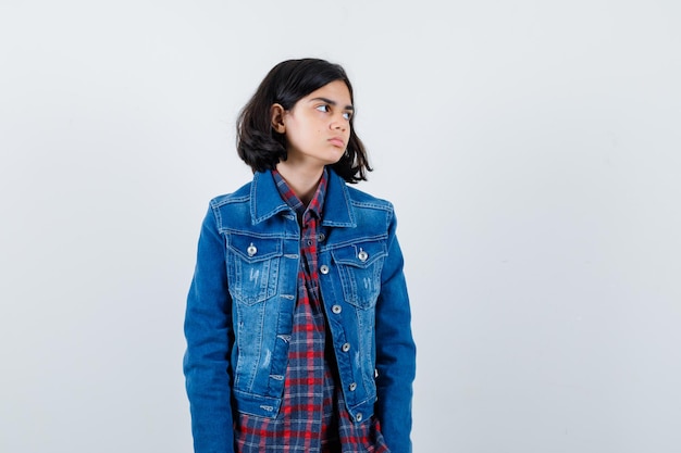 Jeune fille regardant loin tout en se présentant à la caméra en chemise à carreaux et veste en jean et l'air mignon, vue de face.