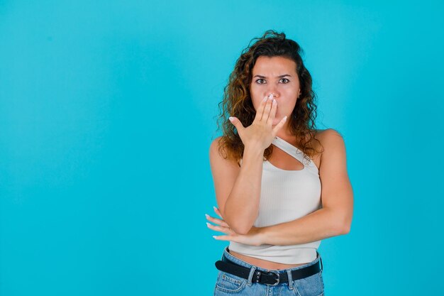 Jeune fille regarde la caméra en tenant la main sur la bouche sur fond bleu