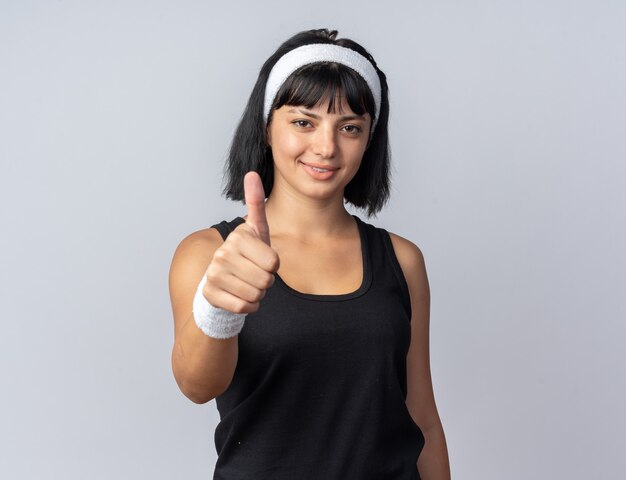 Jeune fille de remise en forme portant un bandeau regardant la caméra souriant confiant montrant le pouce vers le haut debout sur blanc