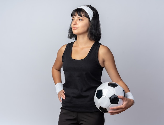 Jeune fille de remise en forme portant un bandeau tenant le football à côté avec un ballon de visage sérieux debout sur blanc