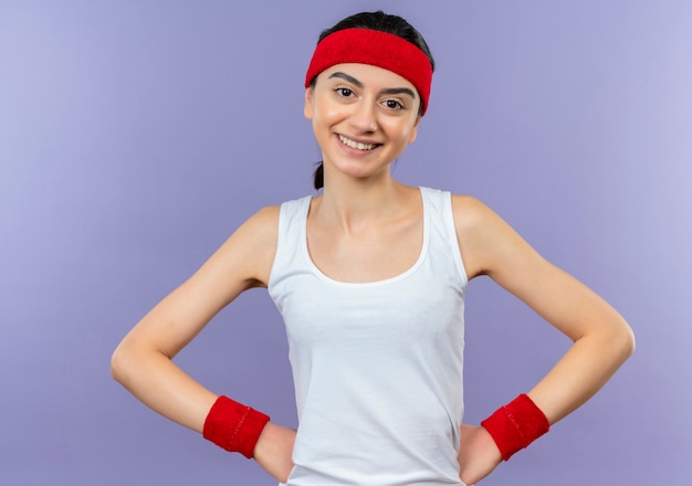 Jeune fille de remise en forme sportswear avec bandeau avec sourire confiant debout sur un mur violet