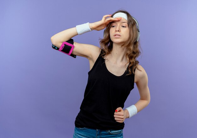 Jeune fille de remise en forme en sportswear noir avec bandeau et brassard de smartphone à la recherche de loin avec la main sur la tête debout sur le mur bleu