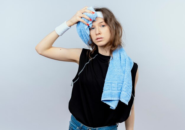 Jeune fille de remise en forme en sportswear noir avec bandeau et casque et serviette sur l'épaule à la fatigue essuyant son front debout sur un mur blanc