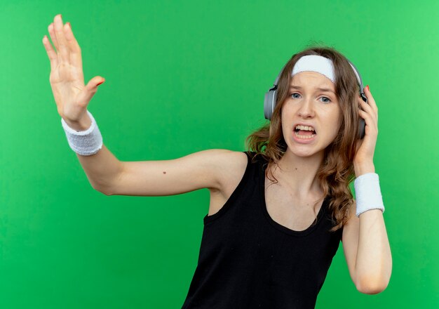 Jeune fille de remise en forme en sportswear noir avec bandeau avec des écouteurs à la confusion et mécontent de faire panneau d'arrêt avec la main debout sur le mur vert