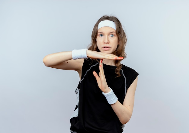 Jeune fille de remise en forme en sportswear noir avec bandeau faisant le geste de temps d'arrêt debout sur un mur blanc