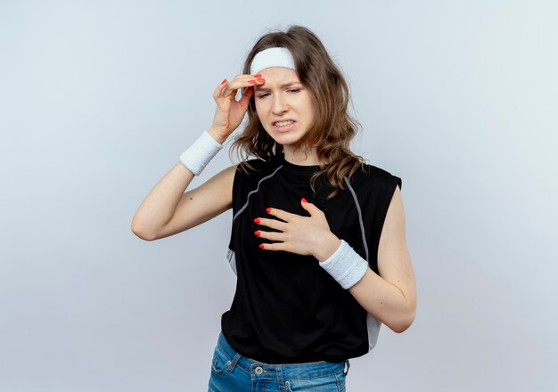 Jeune fille de remise en forme en sportswear noir avec bandeau à mécontent de toucher sa tête ayant de forts maux de tête debout sur un mur blanc