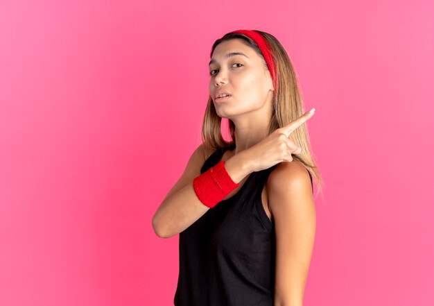 Jeune fille de remise en forme en sportswear noir et bandeau rouge à la confiance pointant vers l'arrière sur rose