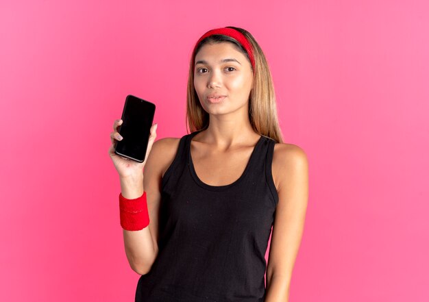 Jeune fille de remise en forme en sportswear noir et bandeau rouge montrant smartphone à la confiance debout sur le mur rose