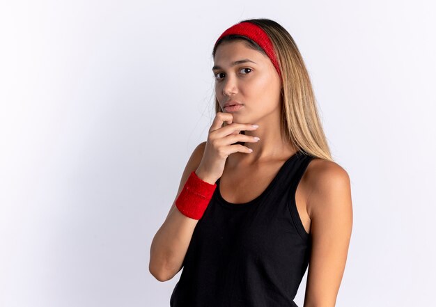 Jeune fille de remise en forme en sportswear noir et bandeau rouge perplexe debout sur un mur blanc