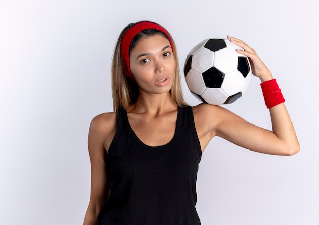 Jeune fille de remise en forme en sportswear noir et bandeau rouge tenant un ballon de football sur l'épaule avec un visage sérieux debout sur un mur blanc