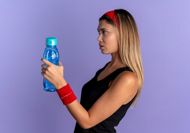 Jeune Fille De Remise En Forme En Sportswear Noir Et Bandeau Rouge Tenant Une Bouteille D'eau à Côté Avec Un Visage Sérieux Sur Bleu