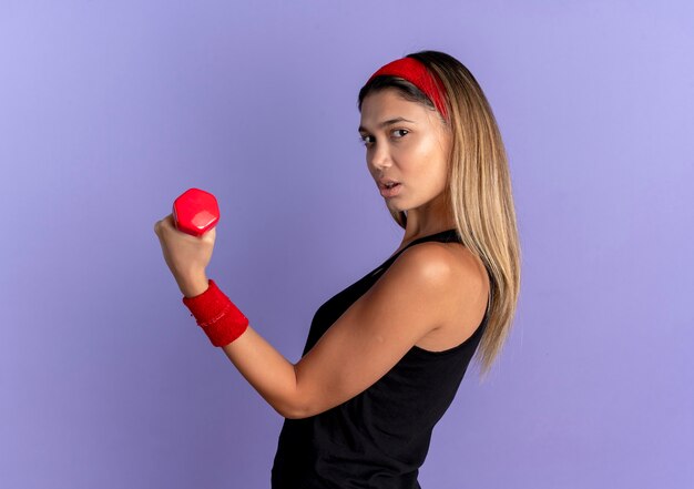 Jeune fille de remise en forme en sportswear noir et bandeau rouge travaillant avec haltère montrant les biceps à la confiance sur le bleu