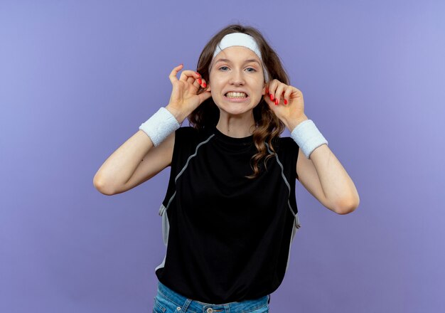 Jeune fille de remise en forme en sportswear noir avec bandeau s'amusant à montrer ses doigts debout sur un mur bleu