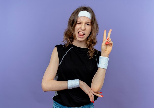 Jeune fille de remise en forme en sportswear noir avec bandeau s'amusant à sortir la langue montrant le signe de la victoire debout sur le mur bleu