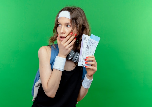Jeune fille de remise en forme en sportswear noir avec bandeau et sac à dos tenant des billets d'avion à côté étonné et surpris debout sur mur vert