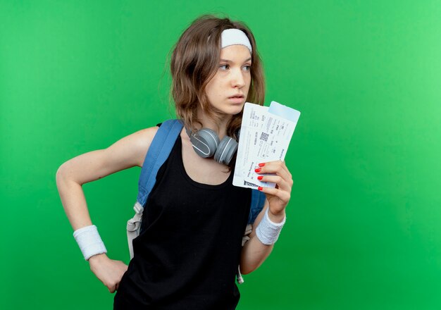 Jeune fille de remise en forme en sportswear noir avec bandeau et sac à dos tenant des billets d'avion à côté avec un visage sérieux debout sur un mur vert