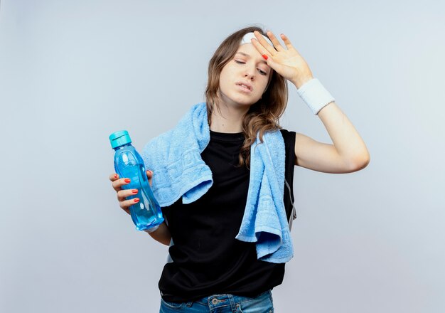 Jeune fille de remise en forme en sportswear noir avec bandeau et serviette autour du cou tenant une bouteille d'eau à la fatigue debout sur un mur blanc