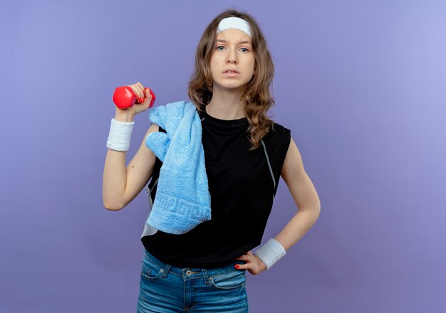 Jeune fille de remise en forme en sportswear noir avec bandeau et serviette sur l'épaule tenant haltère faisant des exercices à la confiance sur le bleu