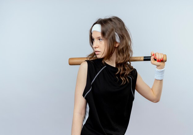 Jeune fille de remise en forme en sportswear noir avec bandeau tenant la chauve-souris basaball à côté avec visage sérieux debout sur un mur blanc