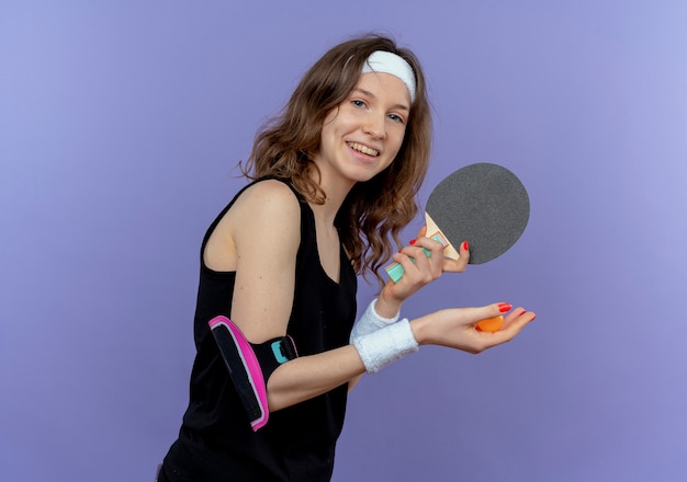 Jeune Fille De Remise En Forme En Sportswear Noir Avec Bandeau Tenant La Raquette Et Les Balles Pour Le Tennis De Table Prêt à Jouer Souriant Joyeusement Debout Sur Le Mur Bleu