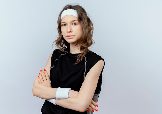 Jeune fille de remise en forme en sportswear noir avec bandeau avec visage sérieux avec les bras croisés debout sur un mur blanc