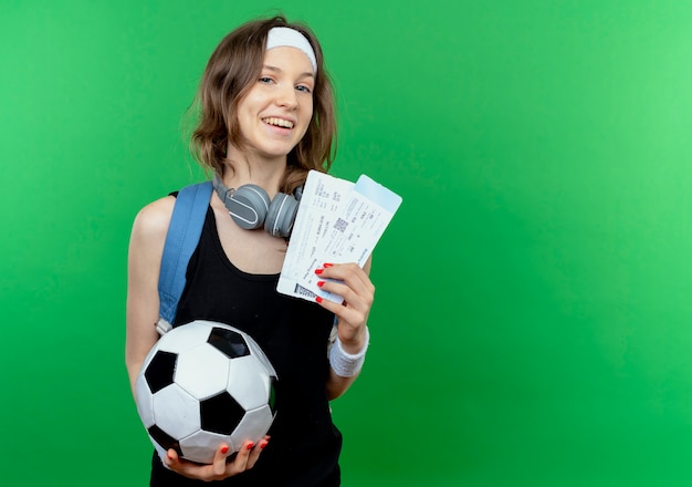 Jeune fille de remise en forme en sportswear noir avec sac à dos et bandeau tenant des billets de football et d'avion souriant joyeusement debout sur un mur vert
