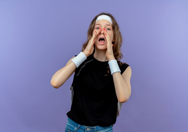 Jeune fille de remise en forme en vêtements de sport noir avec bandeau criant ou appelant avec les mains près de la bouche debout sur le mur bleu