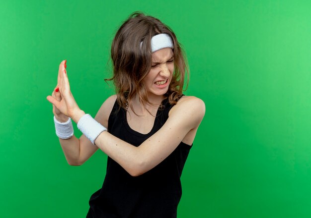 Jeune fille de remise en forme en vêtements de sport noir avec bandeau faisant un geste de défense avec les mains avec les mains comme disant ne pas se rapprocher debout sur le mur vert