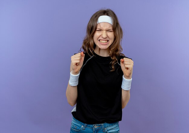 Jeune fille de remise en forme en vêtements de sport noir avec bandeau frustré va sauvage serrant les poings debout sur le mur bleu