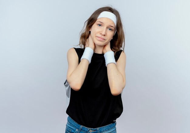 Jeune fille de remise en forme en vêtements de sport noir avec bandeau à la mal de toucher le cou ayant des douleurs debout sur un mur blanc