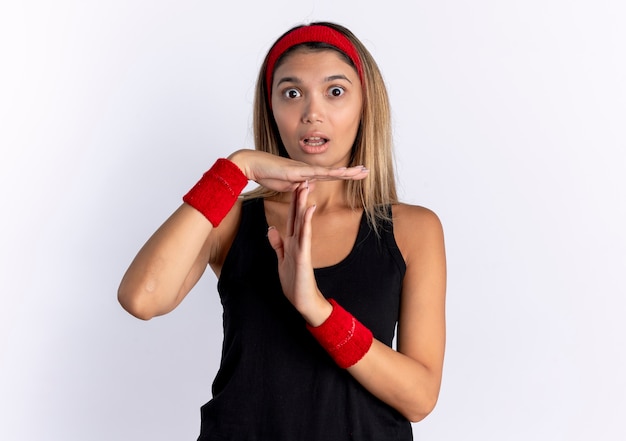 Jeune fille de remise en forme en vêtements de sport noir et bandeau rouge confus faisant le geste de temps avec les mains debout sur un mur blanc