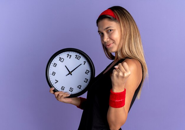 Jeune fille de remise en forme en vêtements de sport noir et bandeau rouge tenant une horloge murale serrant le poing avec un visage sérieux debout sur un mur bleu