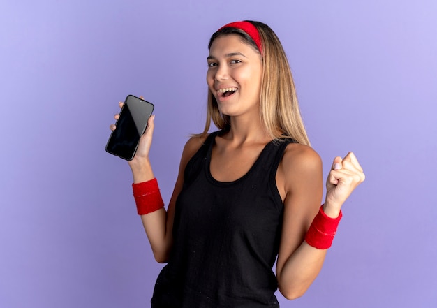 Jeune Fille De Remise En Forme En Vêtements De Sport Noir Et Bandeau Rouge Tenant Le Poing Serrant Le Smartphone Heureux Et Excité Sur Bleu