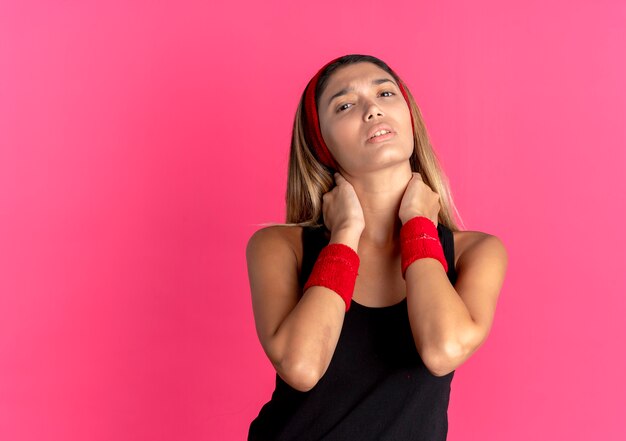 Jeune fille de remise en forme en vêtements de sport noir et bandeau rouge touchant son cou à la douleur ressentie mal debout sur le mur rose