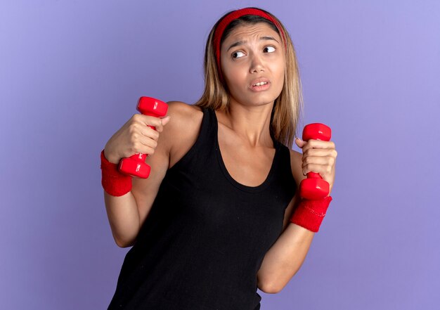 Jeune fille de remise en forme en vêtements de sport noir et bandeau rouge travaillant avec des haltères à la confusion debout sur le mur bleu