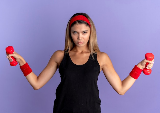 Jeune fille de remise en forme en vêtements de sport noir et bandeau rouge travaillant avec des haltères lookign at camera avec visage sérieux sur bleu