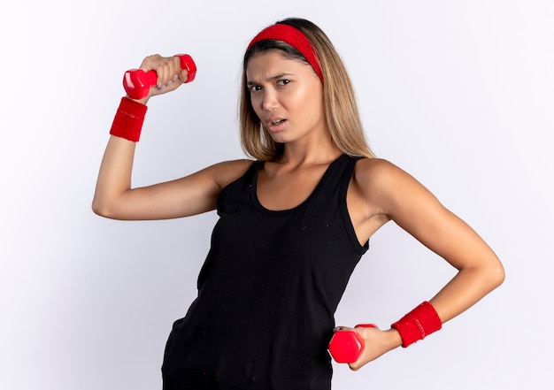 Jeune fille de remise en forme en vêtements de sport noir et bandeau rouge travaillant avec des haltères à la recherche avec un visage sérieux debout sur un mur blanc