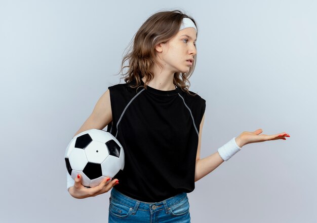 Jeune fille de remise en forme en vêtements de sport noir avec bandeau tenant un ballon de football à côté avec le bras comme demandant debout sur un mur blanc