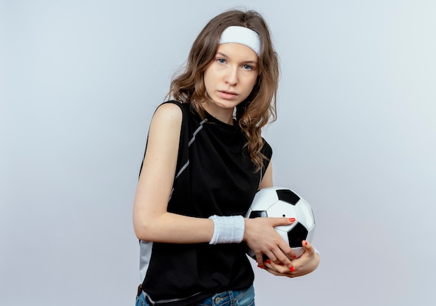 Jeune Fille De Remise En Forme En Vêtements De Sport Noir Avec Bandeau Tenant Un Ballon De Football Mécontent De Se Tenir Debout Sur Un Mur Blanc