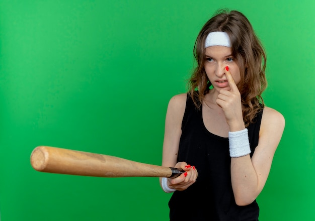 Jeune fille de remise en forme en vêtements de sport noir avec bandeau tenant la chauve-souris basaball regardant quelqu'un qui vous regarde geste pointant avec le doigt sur son œil debout sur le mur vert