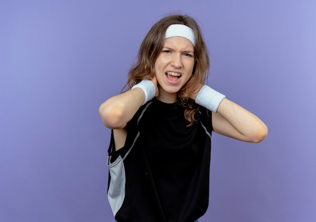 Jeune fille de remise en forme en vêtements de sport noir avec bandeau touchant son cou à mécontent d'avoir la douleur debout sur le mur bleu