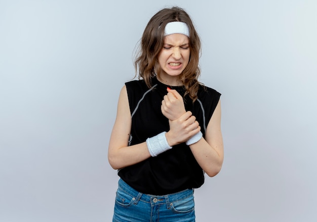 Jeune fille de remise en forme en vêtements de sport noir avec bandeau touchant son poignet à la souffrance souffrant de douleur debout sur un mur blanc