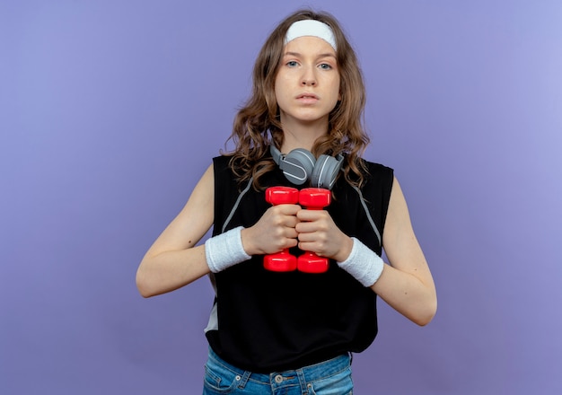 Jeune fille de remise en forme en vêtements de sport noir avec bandeau travaillant avec des haltères à la confiance debout sur le mur bleu