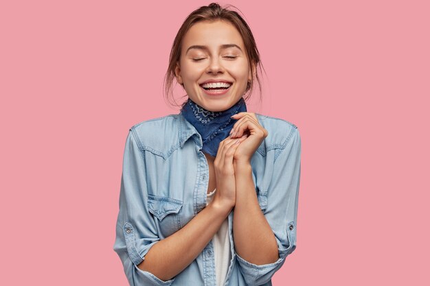 Une jeune fille rêveuse et heureuse garde les yeux fermés, sourit largement, tient les mains ensemble, imagine quelque chose d'agréable à l'esprit